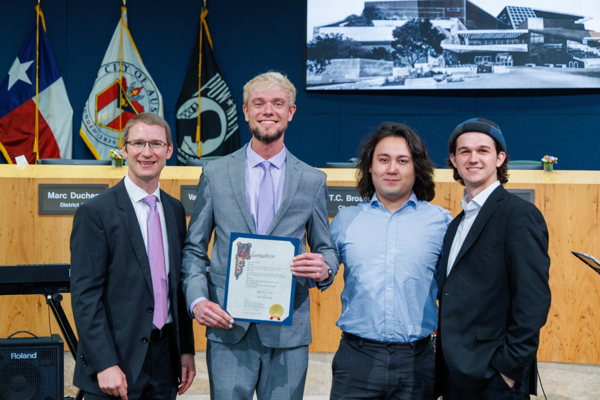 Austin Kimble holding the Austin Kimble Day proclamation beside a city representative and his two fellow musicians at City Hall
